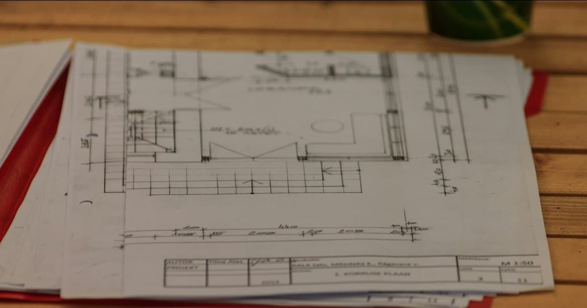 Een stapel bouwtekeningen op een houten tafel. Voordat de calculator een prijs maakt, moet dit allemaal gelezen zijn, samen met het bestek en de nota van inlichtingen.