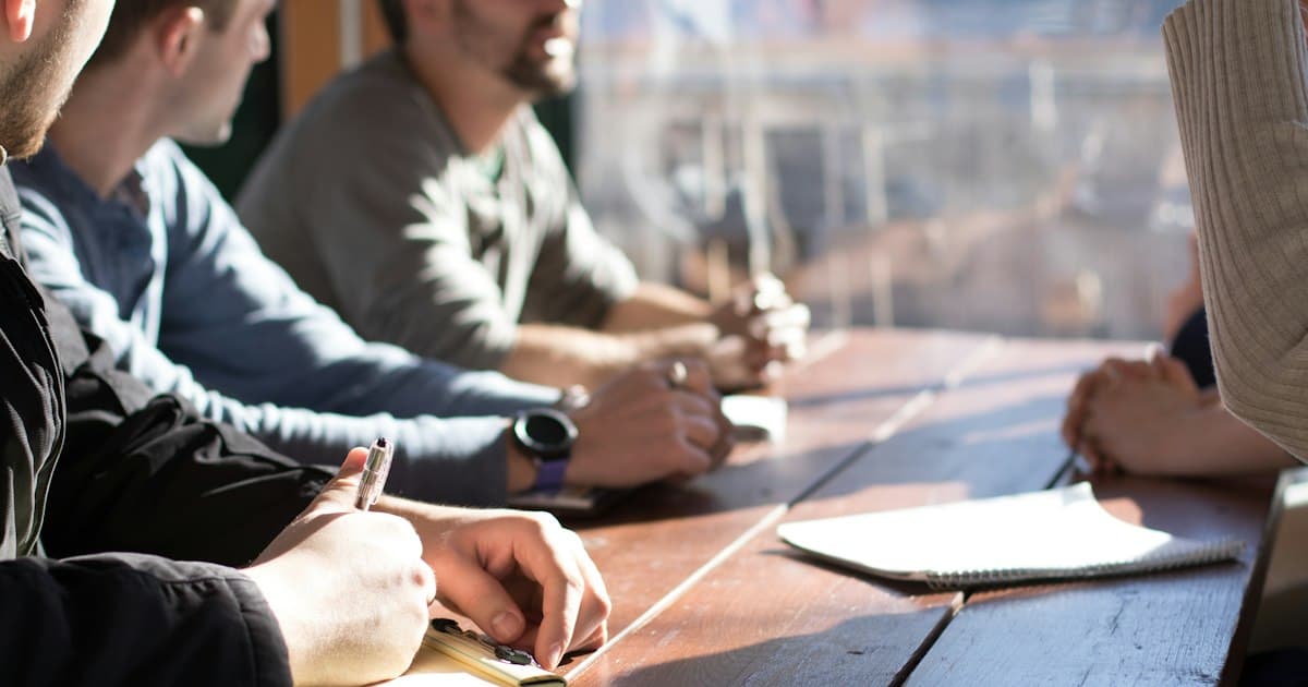 Colleagues around a wooden table with notebooks. The five questions are answered together, in writing.